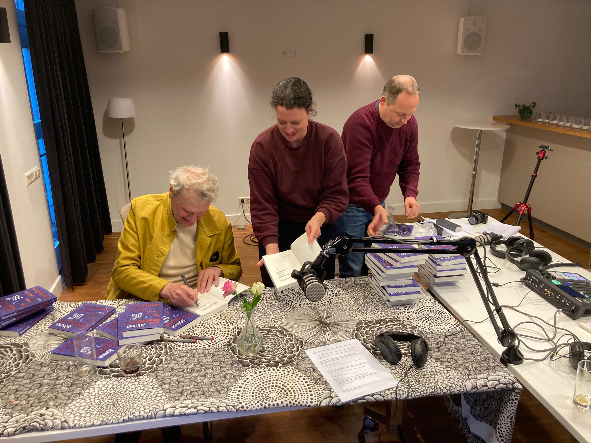 Adriaan van Dis signeert een stapel boeken aan een tafel, geassisteerd door twee mensen van de uitgeverij.